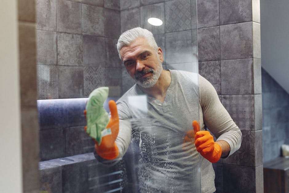 Grey haired male with beard in orange rubber gloves attentively cleaning shower glass in bathroom
