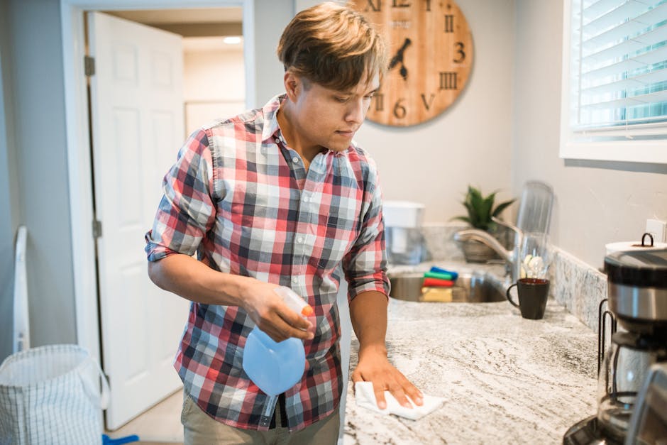 Asian man wiping kitchen counter using a cleaning solution in modern kitchen setting.